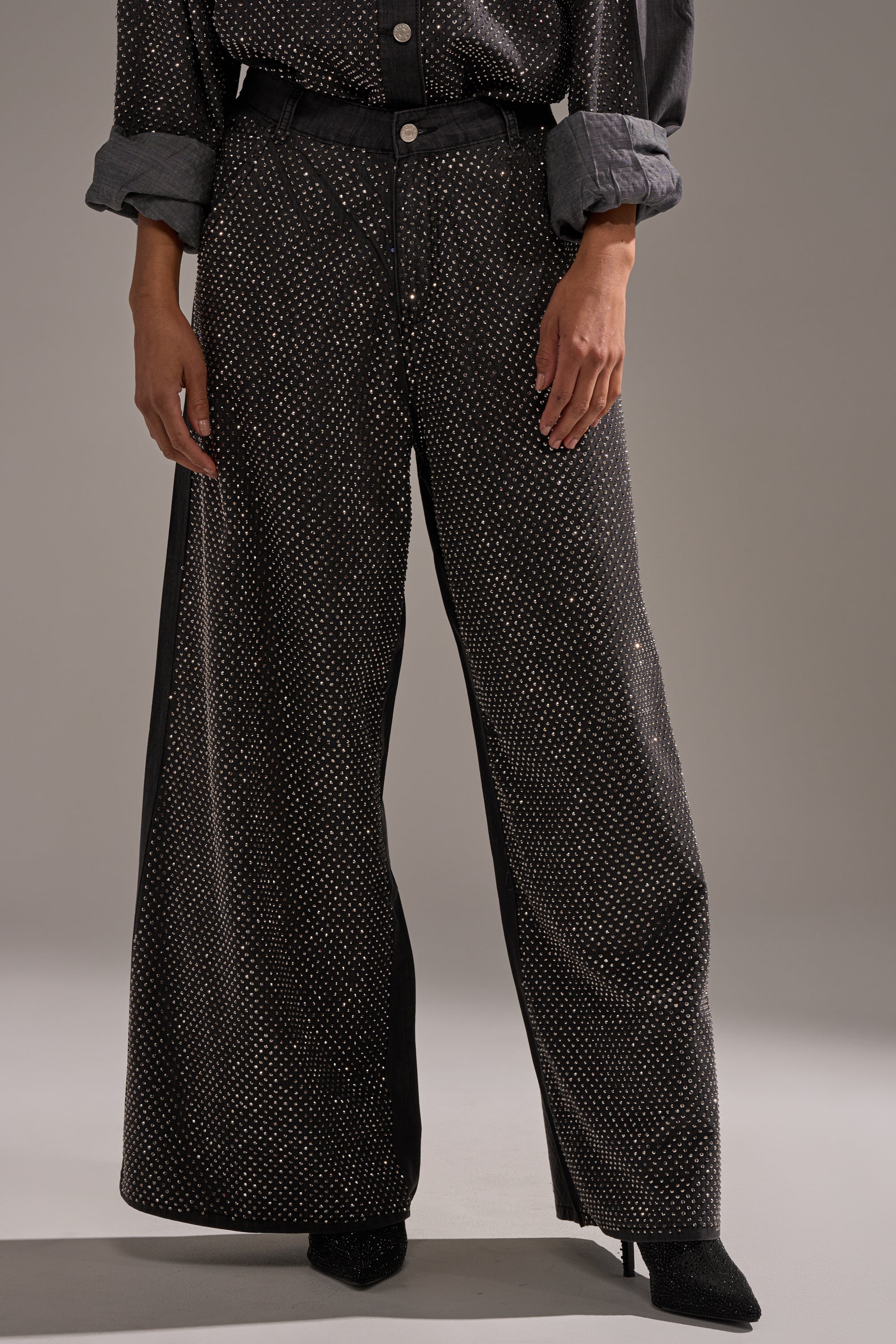 Wearing the BLING BLING DENIM RELAXED PANT IN BLACK—high-waisted, wide-leg denim with small white polka dots—paired with a matching rolled-sleeve top and pointed-toe shoes, standing against a neutral background. 0