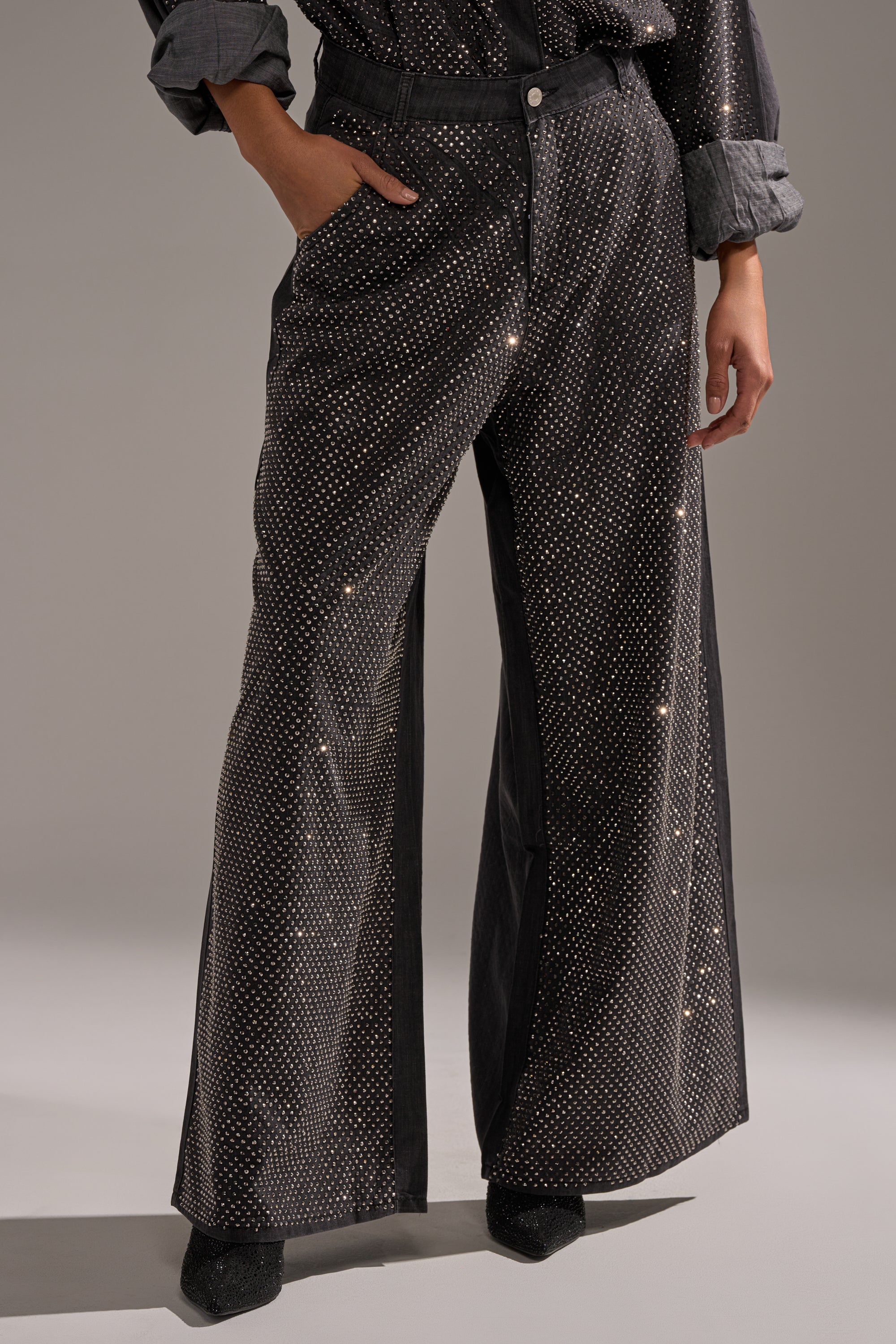 Wearing the BLING BLING DENIM RELAXED PANT IN BLACK adorned with rhinestones, a matching shirt with rolled-up sleeves, and black shoes, a person stands confidently with one hand in their pocket. 1