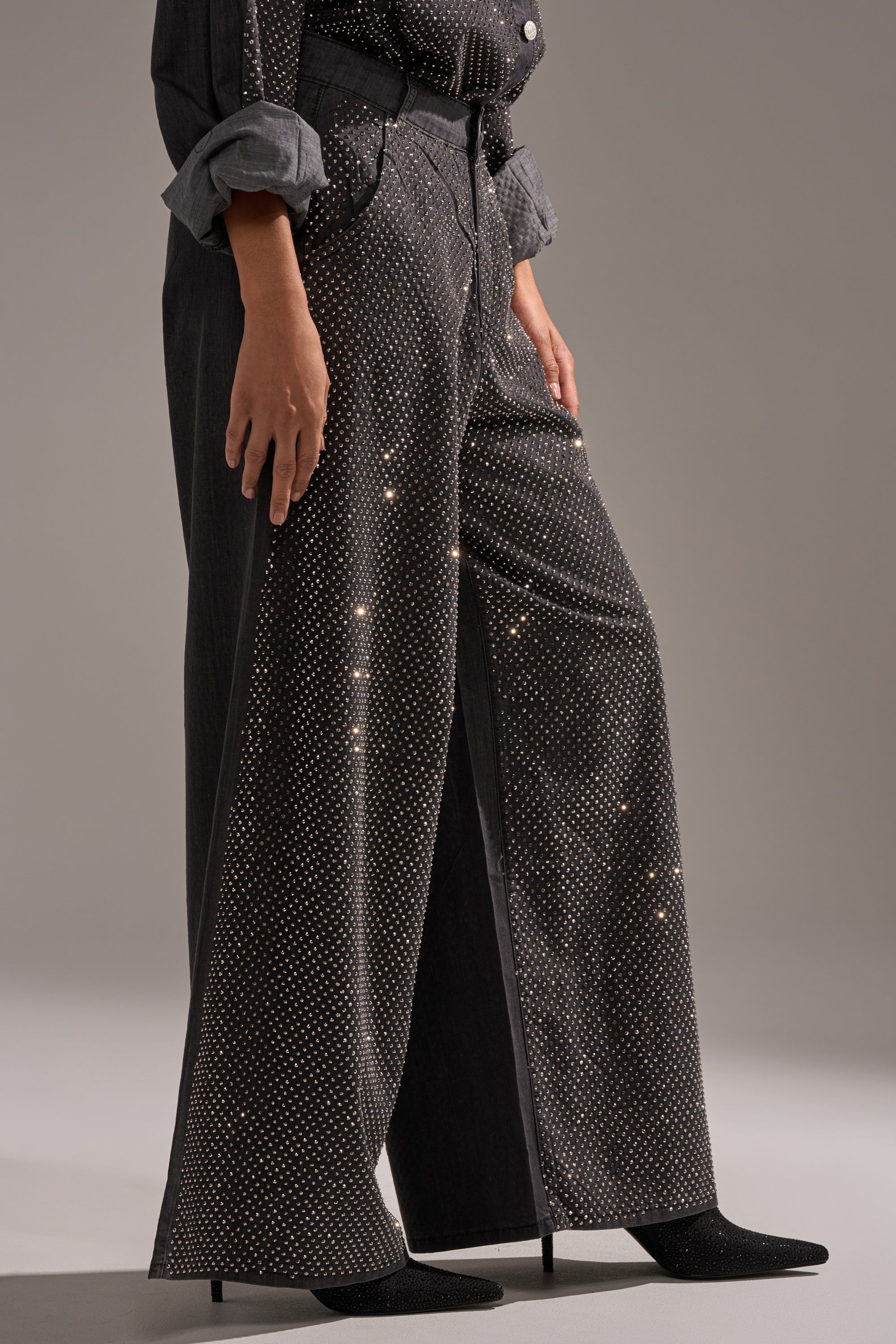 A person models the BLING BLING DENIM RELAXED PANT IN BLACK with a matching shirt and black pointed-toe heeled boots, posing stylishly against a plain background. 2