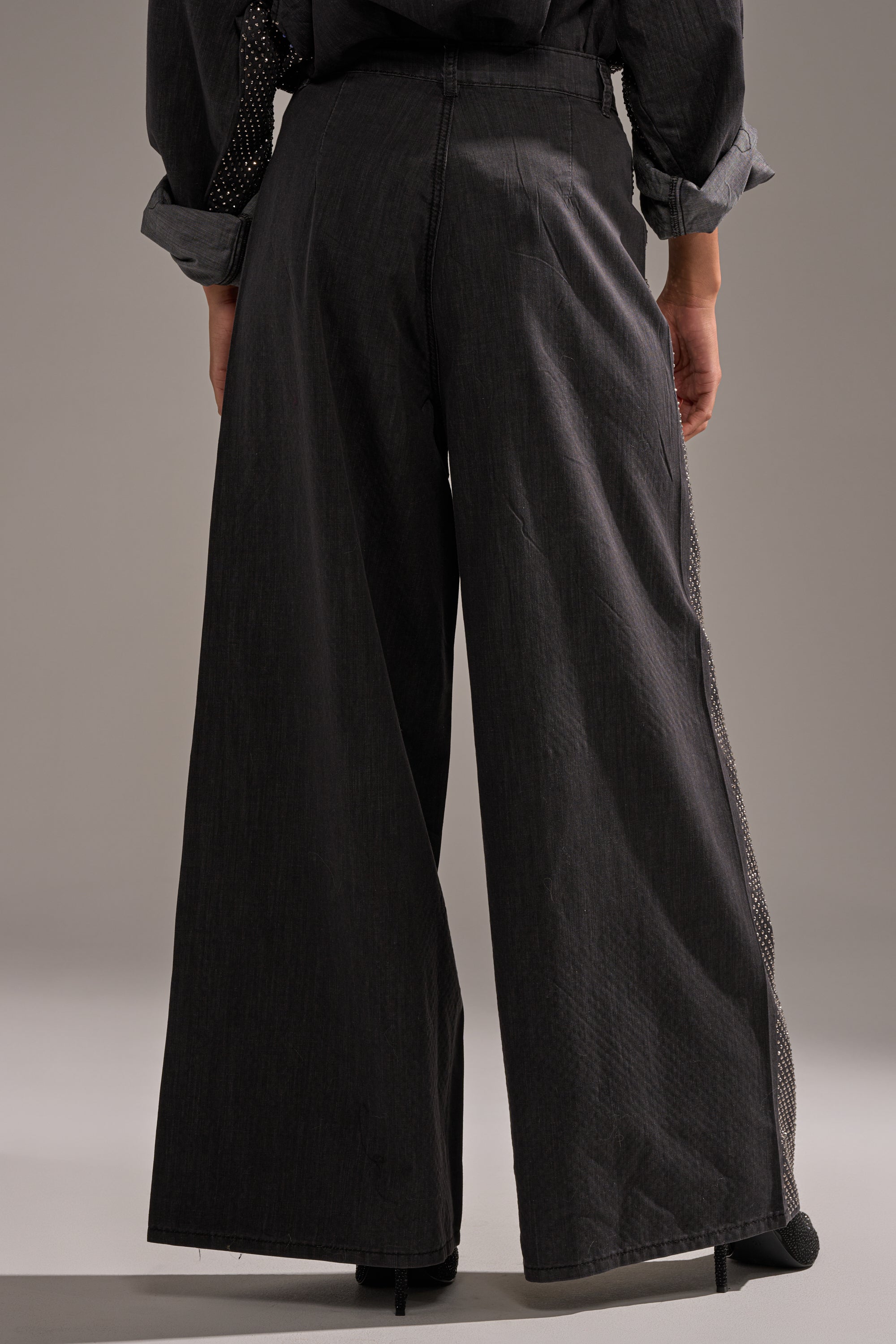 A person wearing BLING BLING DENIM RELAXED PANT IN BLACK and black heeled shoes is shown from the back against a plain background. 3