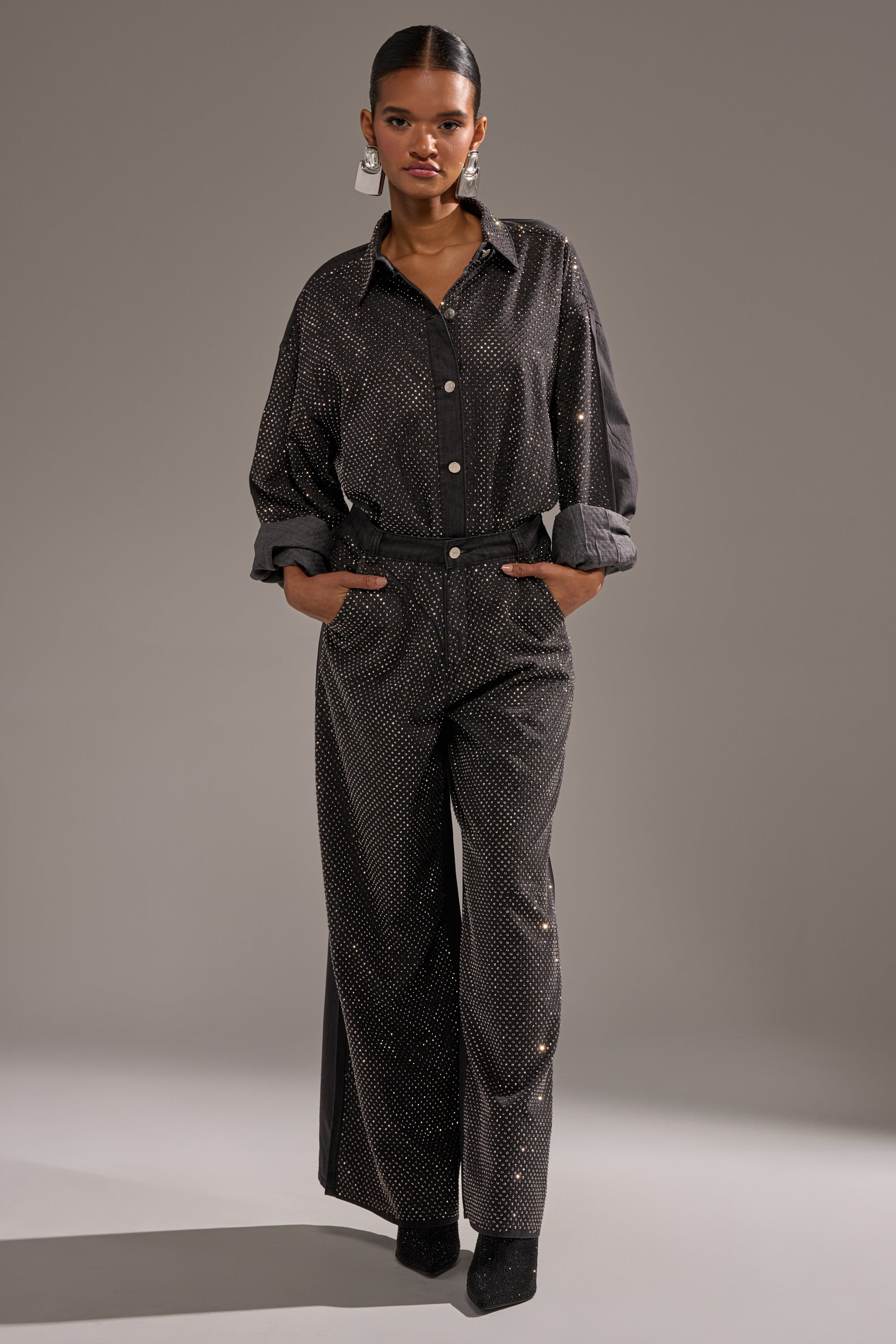 A woman stands with hands in pockets, wearing the BLING BLING DENIM RELAXED PANT IN BLACK featuring subtle sparkling dots. She pairs them with a rolled-sleeve top, pointed black shoes, statement earrings, and has her hair pulled back. 4