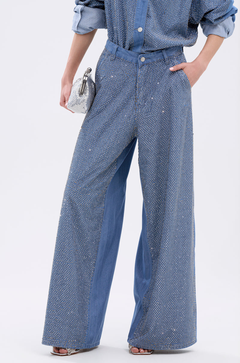 A person wears the BLING BLING DENIM RELAXED PANT—high-rise, wide-leg jeans with a subtle shimmer—paired with a matching blue shirt, holding a small silver clutch, standing against a simple light background.