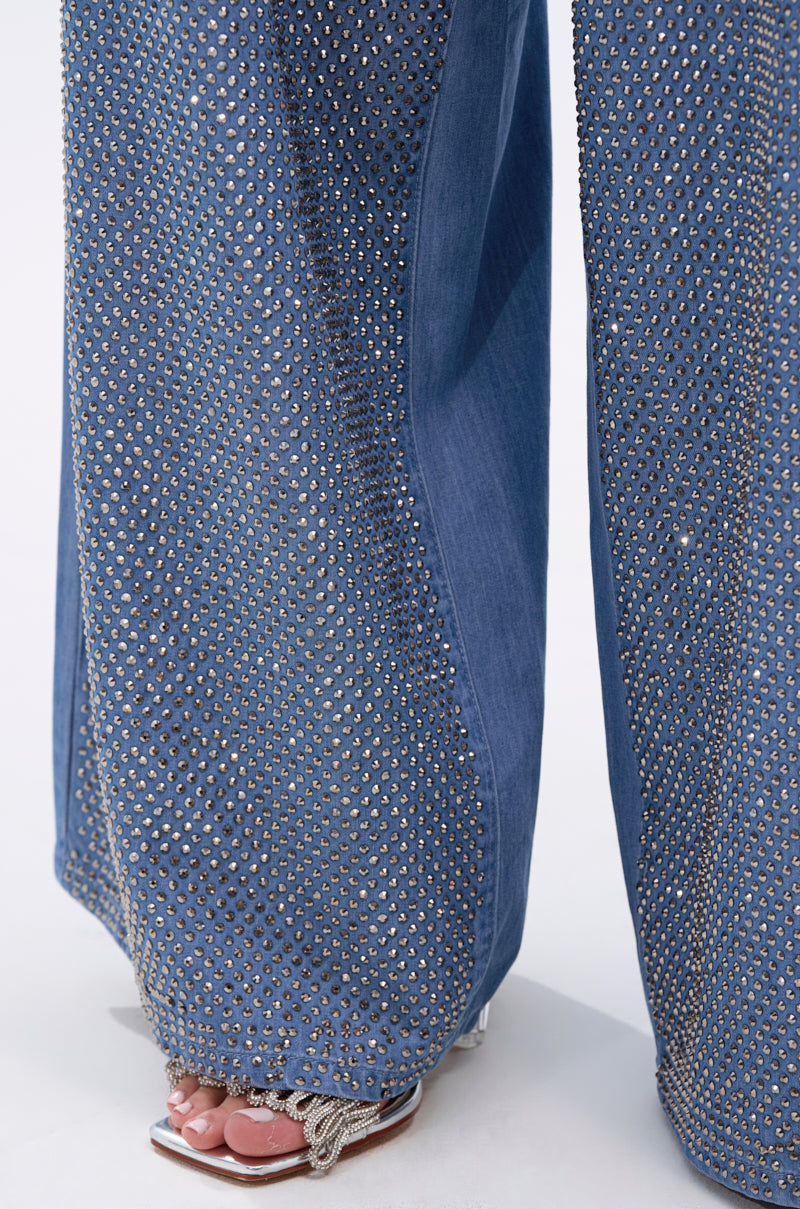 A close-up of the BLING BLING DENIM RELAXED PANT, a high-rise, wide-leg denim adorned with silver studs, styled with open-toed sandals featuring silver chain details across the toes.