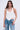 A woman with long wavy hair models the DESTINY CORSET, a white halter-top with halter straps, paired with light blue zipper-detailed jeans against a plain light background.