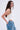 A woman with long wavy brown hair models the DESTINY CORSET, a white sleeveless halter top, paired with ripped blue jeans accented by metal key details, standing sideways and looking at the camera against a plain light background.