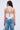 A woman with wavy brown hair is seen from the back wearing the DESTINY CORSET, a white halter top with a lace-up back, paired with faded distressed blue jeans, and holding a small chain-strap bag.