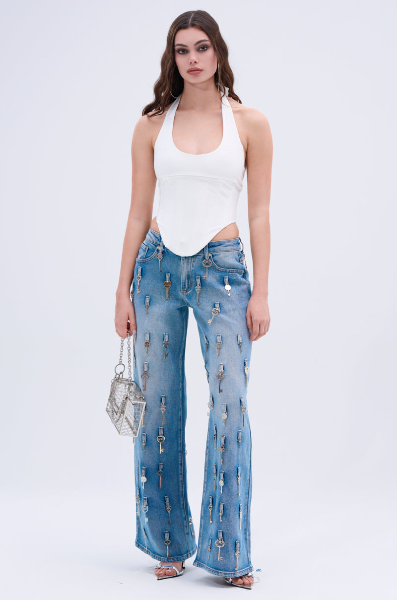 A woman stands against a plain background wearing the DESTINY CORSET—a white halter top with halter-style straps—and light blue flared jeans adorned with metal keys, paired with a small silver handbag and silver open-toe heels.