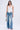 A woman stands against a plain background wearing the DESTINY CORSET—a white halter top with halter-style straps—and light blue flared jeans adorned with metal keys, paired with a small silver handbag and silver open-toe heels.