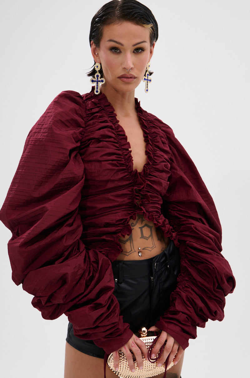 A woman with slicked-back hair models the IN IT TO WIN IT RUCHED BLOUSE IN BURGUNDY featuring voluminous sleeves and a plunging neckline, styled with black shorts, cross earrings, a gold clutch, and visible tattoos against a white background.