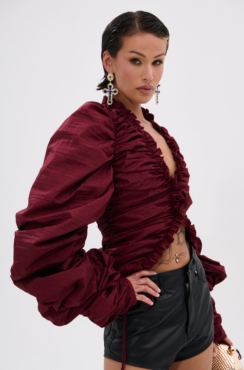 A person poses confidently in the IN IT TO WIN IT RUCHED BLOUSE IN BURGUNDY, featuring a plunging neckline and puffed sleeves, paired with black high-waisted shorts. Slicked-back hair, cross earrings, and chest tattoos finish the bold style.