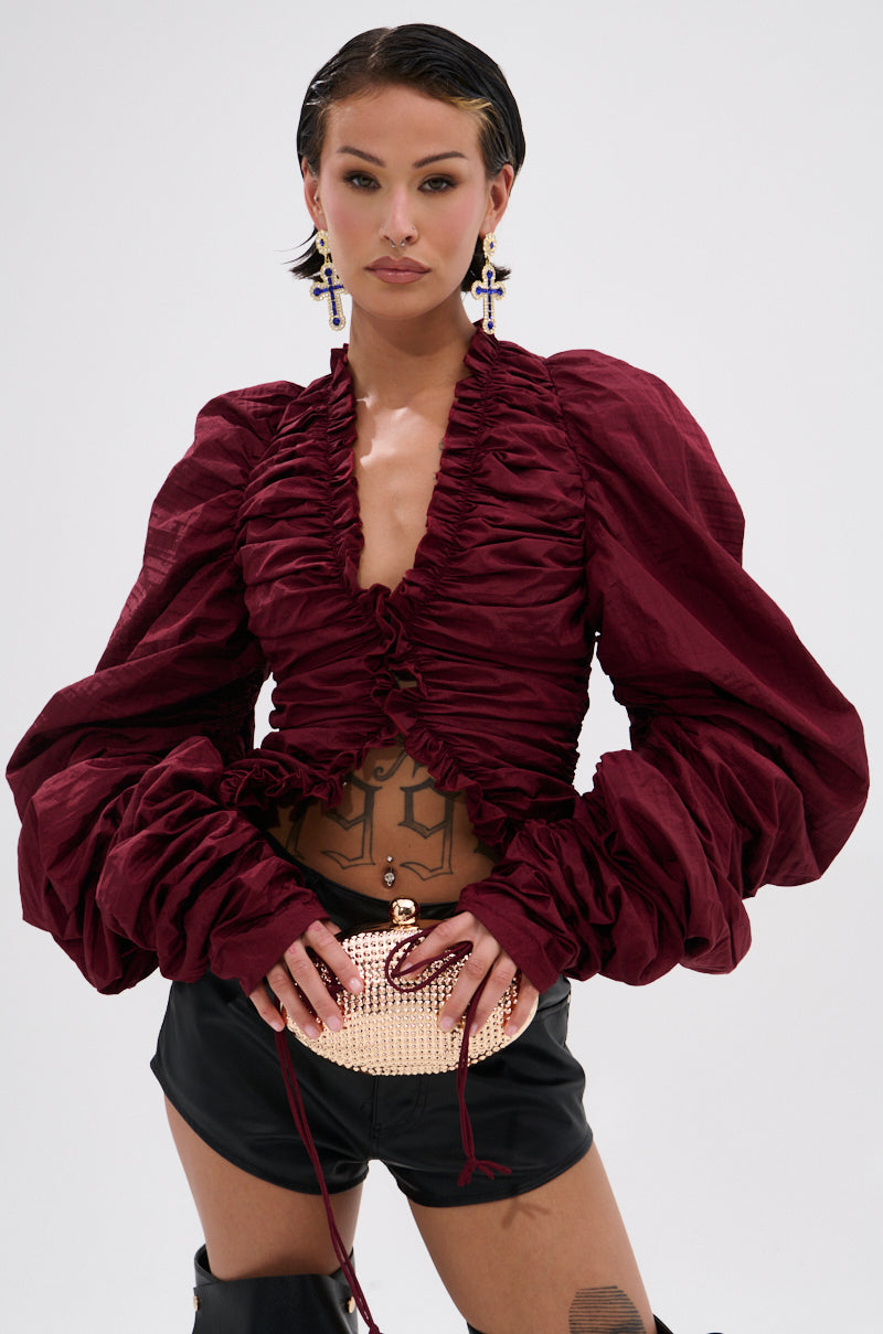 A woman with short dark hair wears the IN IT TO WIN IT RUCHED BLOUSE IN BURGUNDY, styled with black shorts, statement earrings, a small gold jeweled clutch, and visible tattoos that highlight her confident look.
