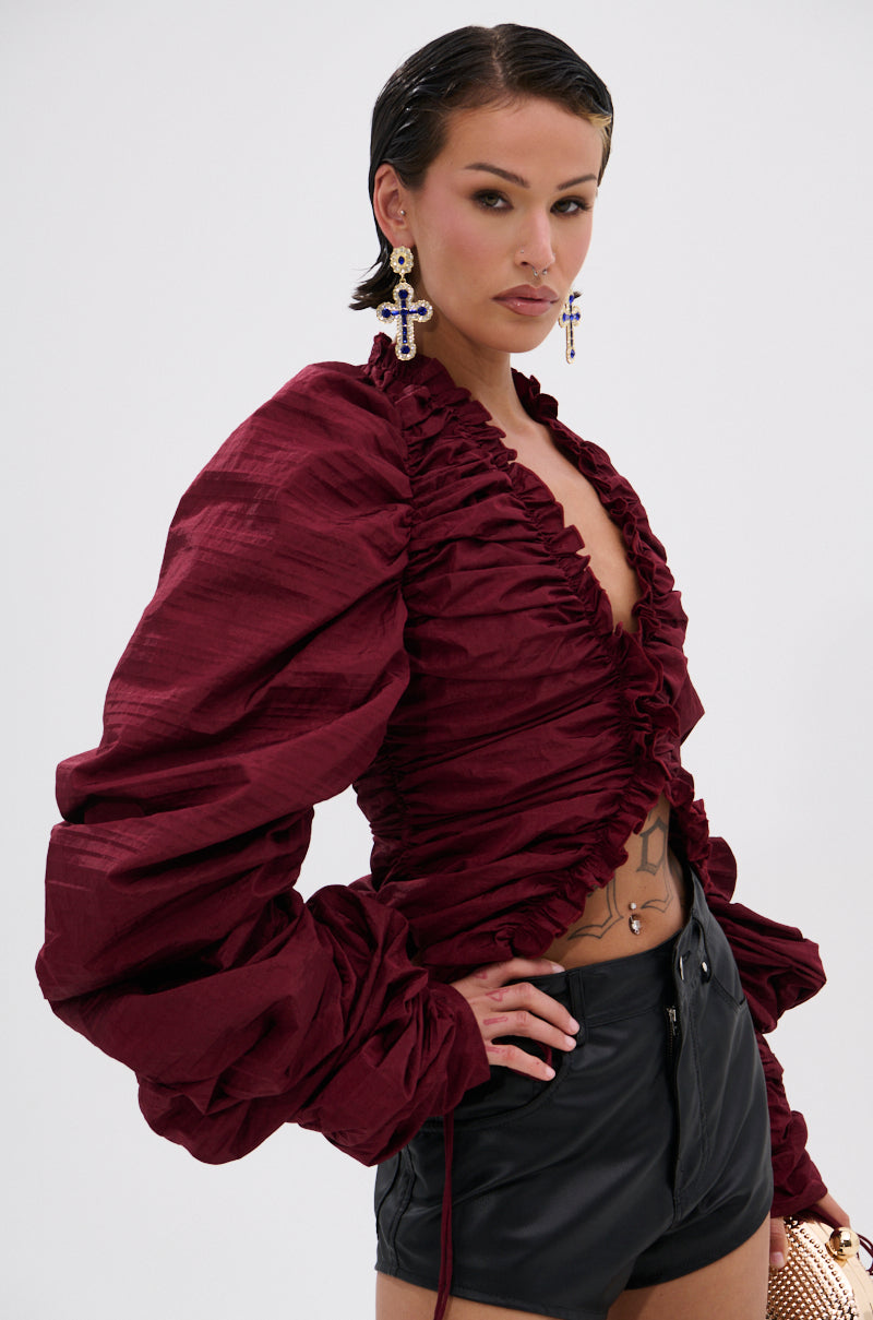 A woman with slicked-back hair wears large cross earrings and the IN IT TO WIN IT RUCHED BLOUSE IN BURGUNDY with a plunging neckline and voluminous sleeves, paired with high-waisted black shorts, posing confidently against a plain light background.