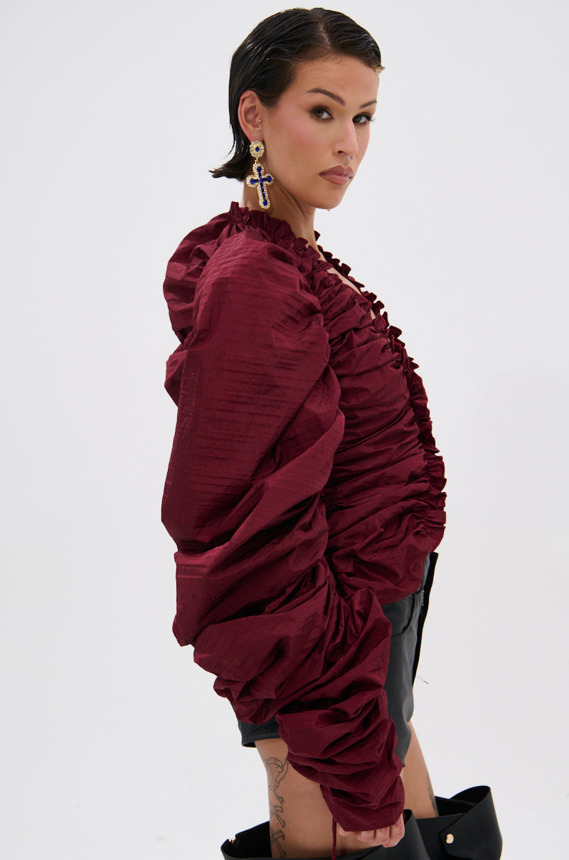 A person with short dark hair poses confidently against a white background, wearing the IN IT TO WIN IT RUCHED BLOUSE IN BURGUNDY with voluminous sleeves, black shorts, and a bold cross earring.