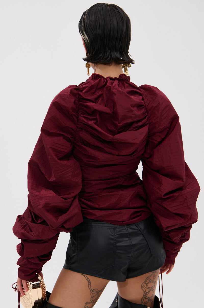 A person with short dark hair is seen from behind wearing the IN IT TO WIN IT RUCHED BLOUSE IN BURGUNDY and black leather shorts. Thigh tattoos are visible as they hold a metallic handbag.