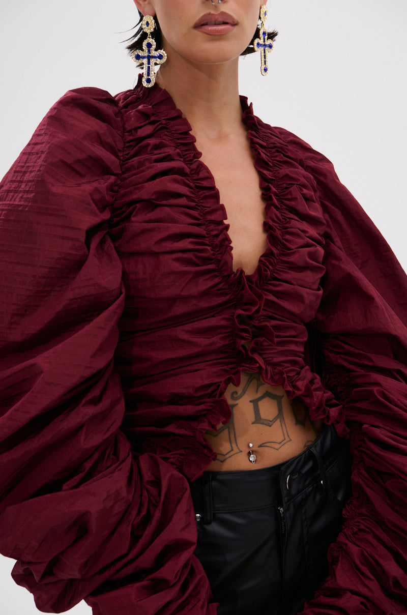 A person wears the IN IT TO WIN IT RUCHED BLOUSE IN BURGUNDY with a plunging neckline, voluminous sleeves, black pants, large cross earrings, and a geometric torso tattoo. Their hair is short and dark.