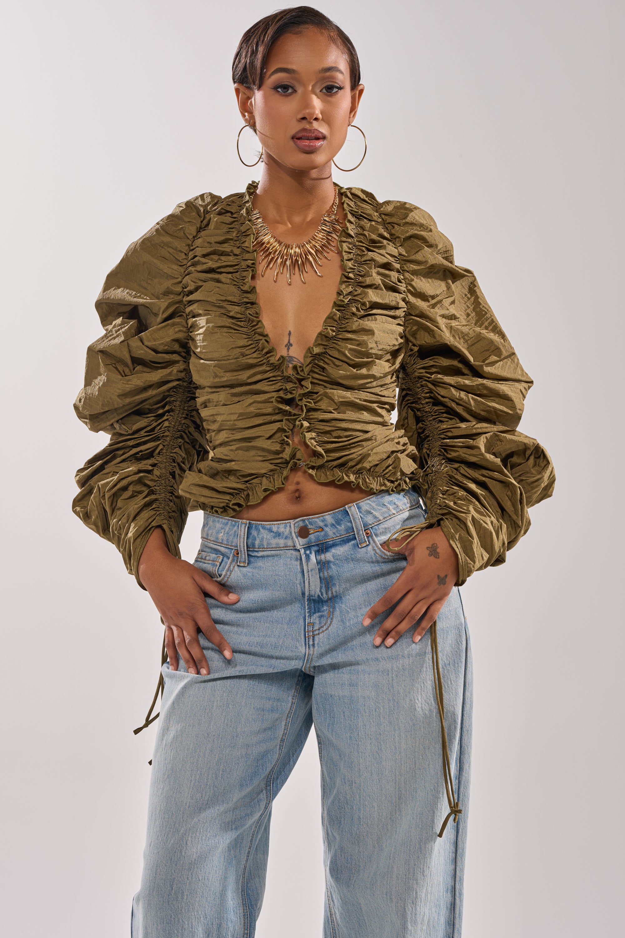 A woman wears the IN IT TO WIN IT RUCHED BLOUSE in olive green, featuring a plunging neckline and voluminous long sleeves, paired with light blue jeans. She accessorizes with large hoop earrings, a gold statement necklace, and has short hair. 1