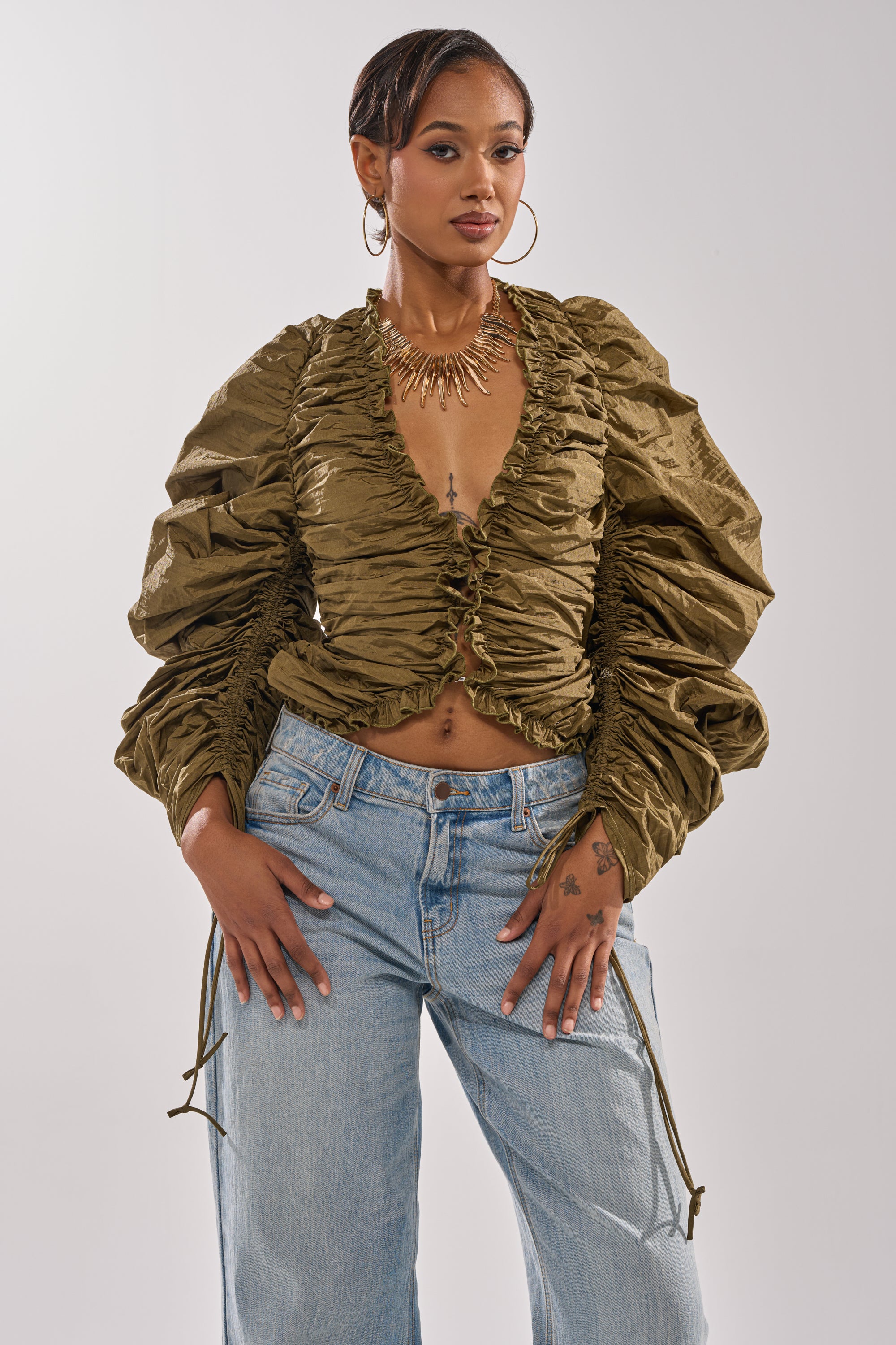 A woman poses confidently in the IN IT TO WIN IT RUCHED BLOUSE, a shiny olive green top featuring voluminous long sleeves and a plunging neckline. She completes her look with a statement necklace, large hoop earrings, and one hand on her hip. 2