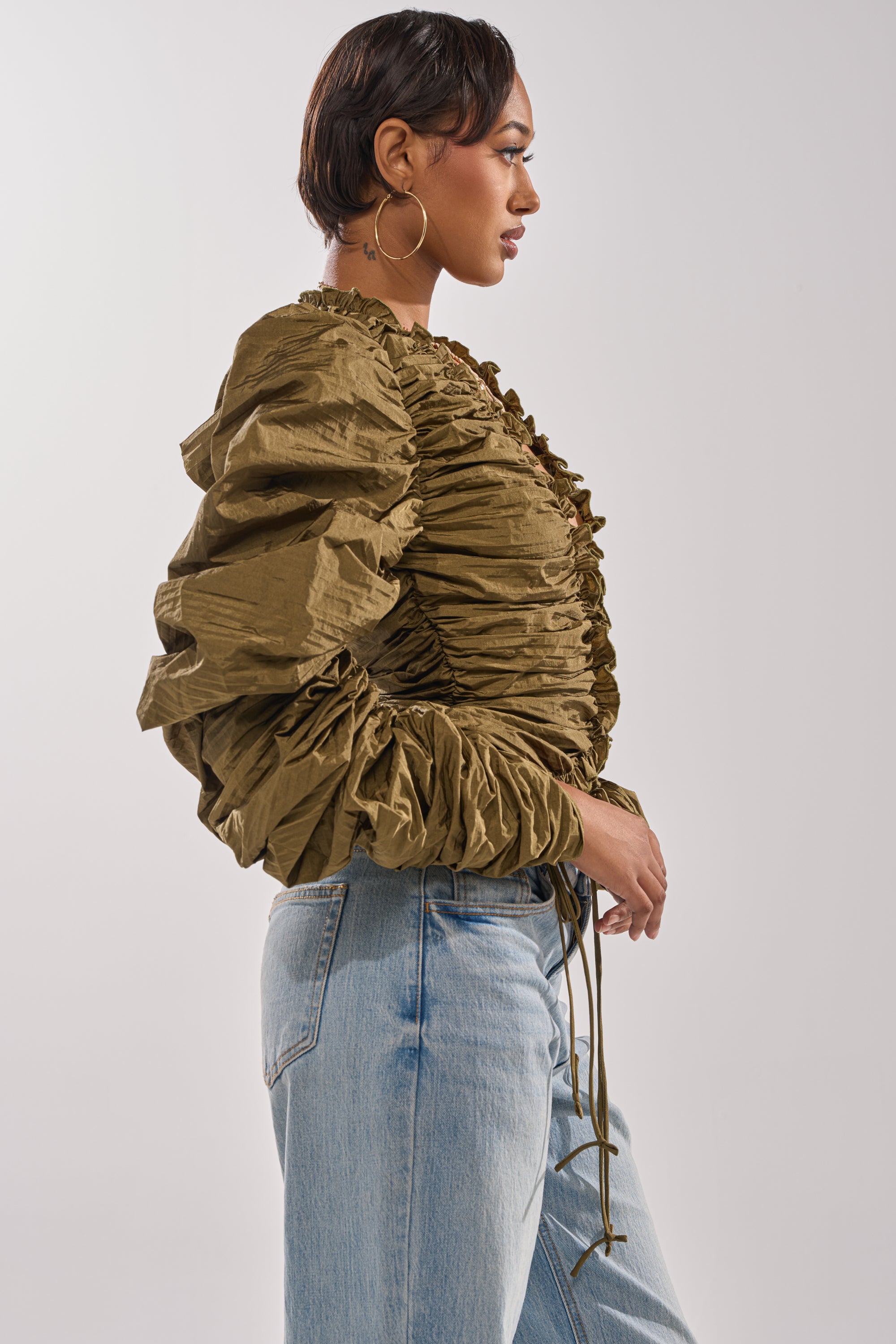A woman with short hair wears large hoop earrings, the IN IT TO WIN IT RUCHED BLOUSE in olive-green with a plunging neckline, and light blue high-waisted jeans, standing in profile against a plain light background. 3