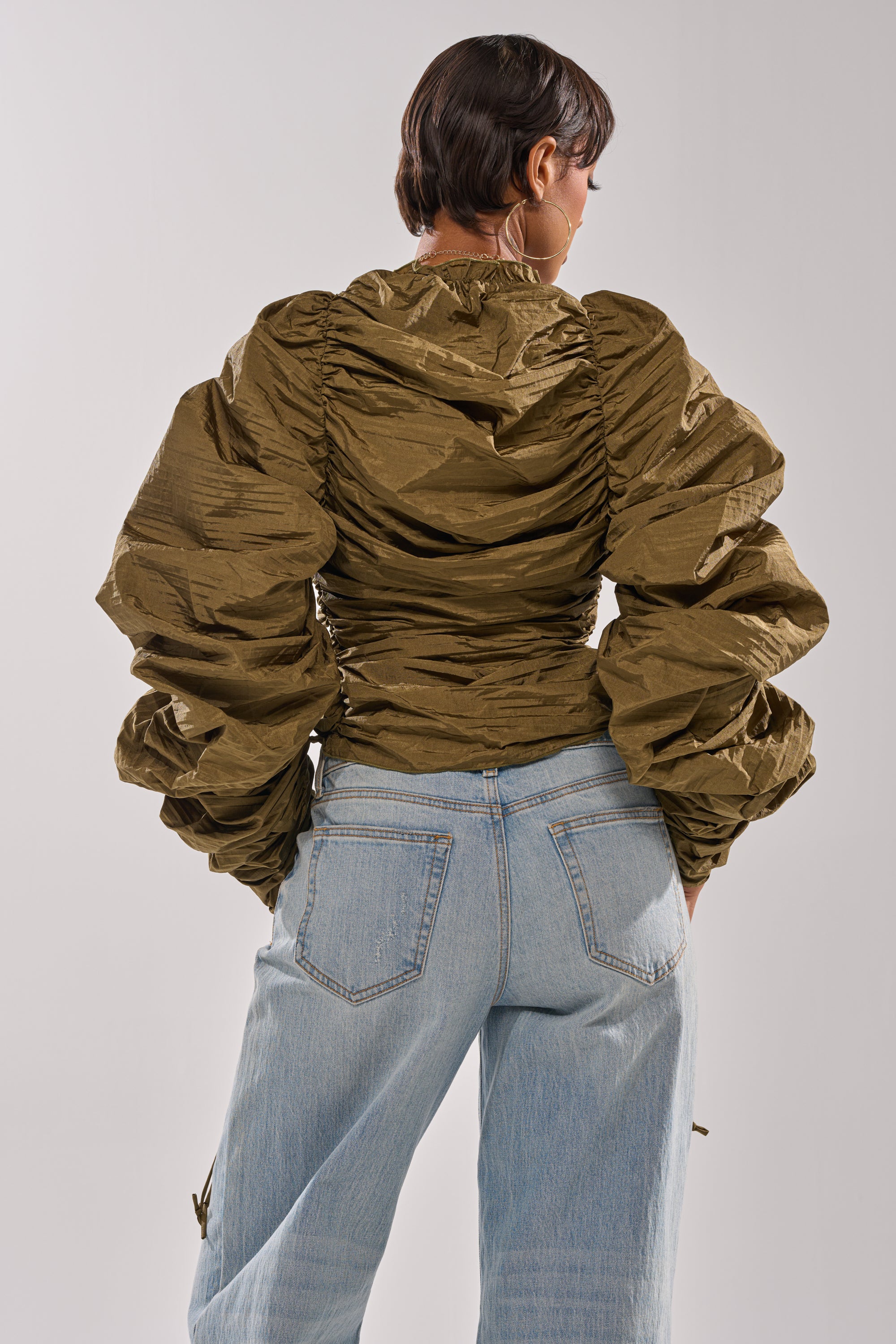 Seen from the back, a person with short hair wears the IN IT TO WIN IT RUCHED BLOUSE in textured olive green with voluminous sleeves, paired with light blue high-waisted jeans against a plain light background. 4