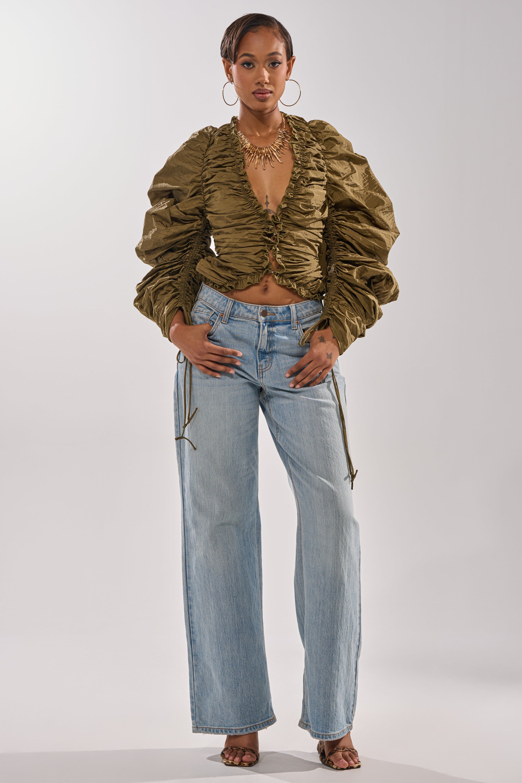 A woman faces forward in the IN IT TO WIN IT RUCHED BLOUSE, a metallic olive green top with adjustable drawstring sleeves, paired with light blue wide-leg jeans, heeled sandals, large hoop earrings, and a statement necklace on a plain white background. 5