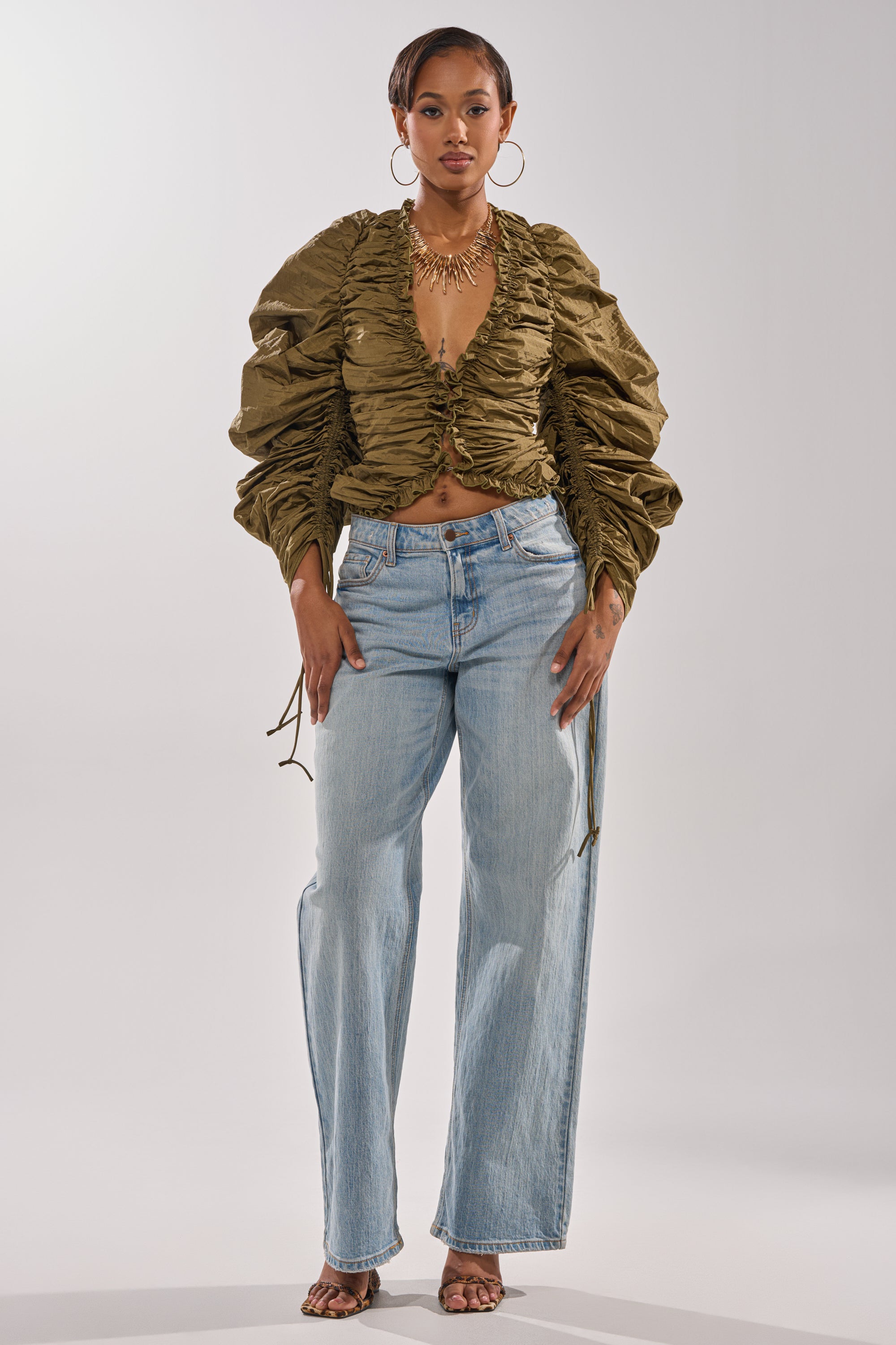 A woman faces forward wearing the IN IT TO WIN IT RUCHED BLOUSE in olive green with adjustable drawstring sleeves, paired with light blue high-waisted jeans, hoop earrings, a gold necklace, and open-toe heels against a light background. 6