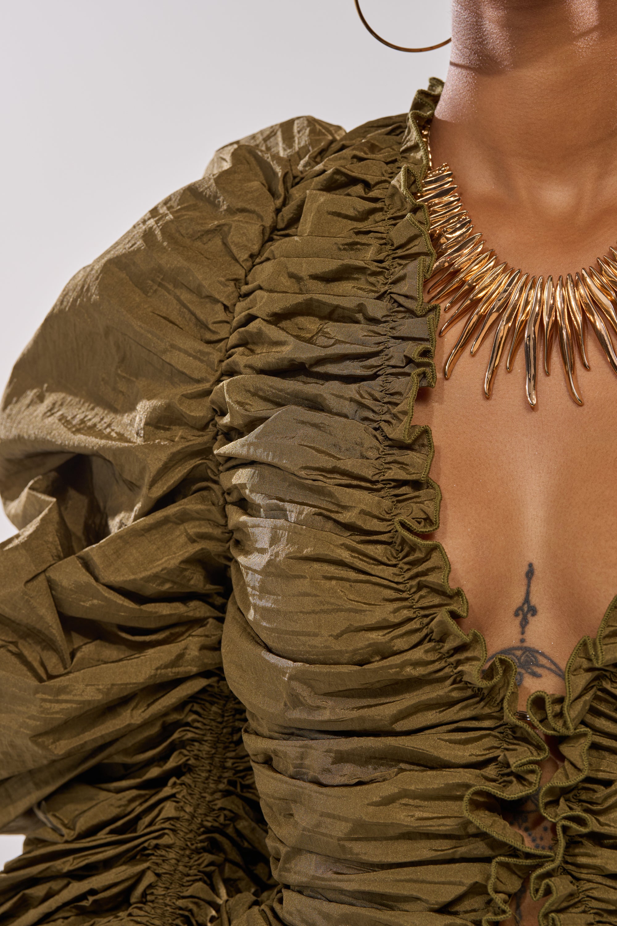 Close-up of a person wearing the IN IT TO WIN IT RUCHED BLOUSE with adjustable drawstring sleeves, paired with a gold spiked necklace. The tattoo on their chest is visible; only the lower face and neck appear. 7
