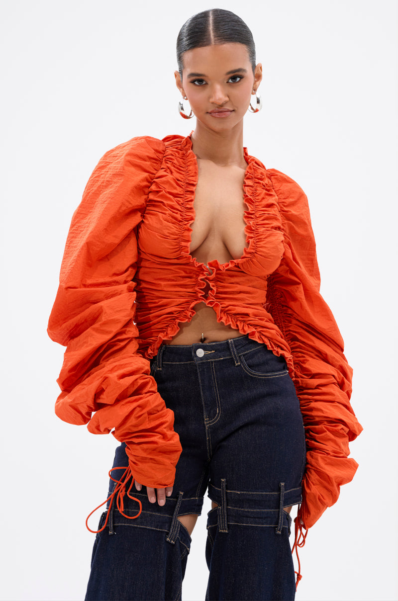 Wearing the IN IT TO WIN IT RUCHED BLOUSE in vibrant orange with a deep V-neck and voluminous sleeves, she pairs it with dark high-waisted jeans featuring cut-outs and ties, standing against a plain background. 0