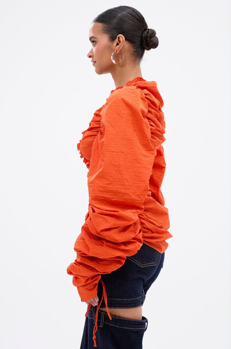 A person stands in profile against a plain background, wearing the vibrant orange IN IT TO WIN IT RUCHED BLOUSE with long sleeves and dark blue jeans. Their hair is tied in a sleek bun, accessorized with large hoop earrings. 2