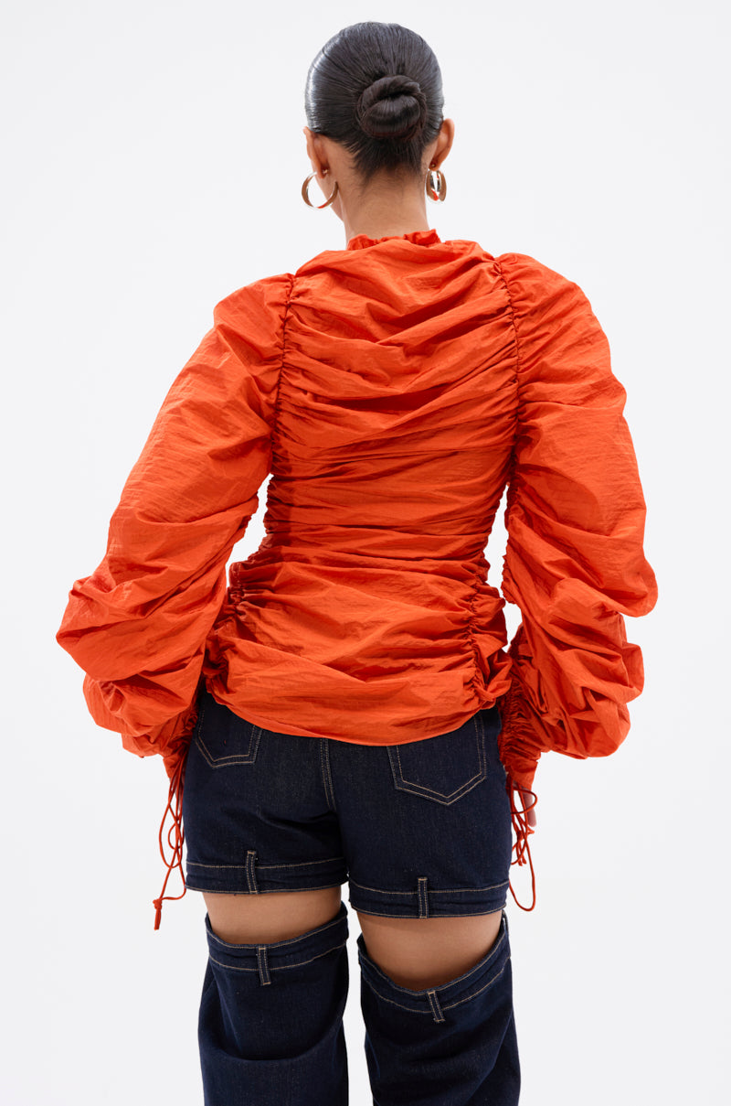 A person with hair in a bun is shown from the back wearing the IN IT TO WIN IT RUCHED BLOUSE in vibrant orange with voluminous sleeves, paired with dark blue jeans featuring thigh cut-outs. 3