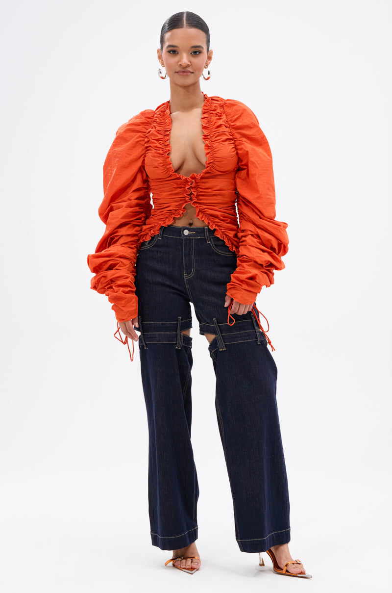 A woman faces forward in the IN IT TO WIN IT RUCHED BLOUSE, a vibrant orange top with a deep plunge, paired with wide-leg dark blue jeans with strap details. She sports slicked-back hair and heeled sandals. 4
