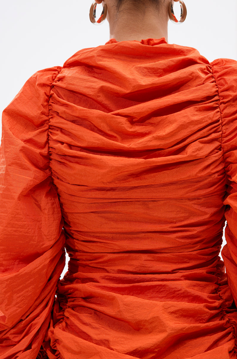 A person is seen from the back wearing the IN IT TO WIN IT RUCHED BLOUSE in vibrant orange, featuring long sleeves and a textured design. Large hoop earrings complete the look against a plain, light-colored background. 6