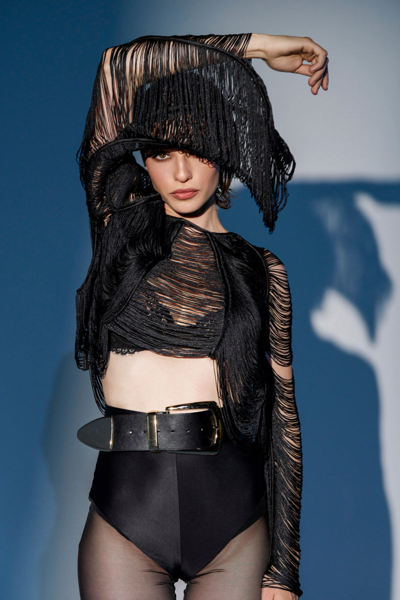 A model poses in the LUCY FRINGE LONG SLEEVE BLOUSE over a black bodysuit, styled with a wide gold-buckle belt and matching fringe hat, raising one arm against a blue background. 1