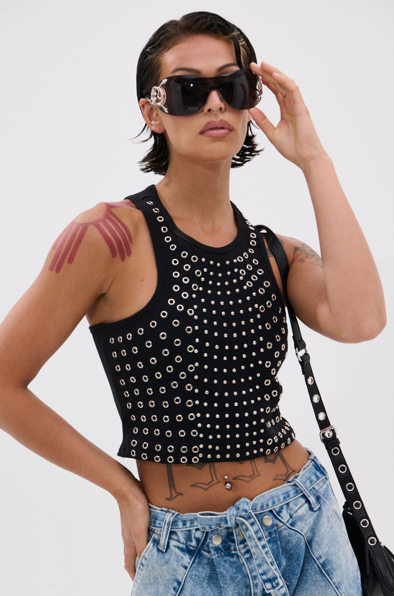 A person with short dark hair and tattoos wears large black sunglasses, acid-wash jeans, and the JANE GROMMET TANK TOP with metallic studs. They pose confidently, holding a hand near their sunglasses. 1