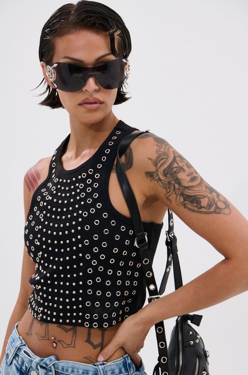 A person with short dark hair and visible arm tattoos wears large sunglasses, blue jeans, a black shoulder bag, and a JANE GROMMET TANK TOP featuring metallic stud details, showing a confident expression. 2