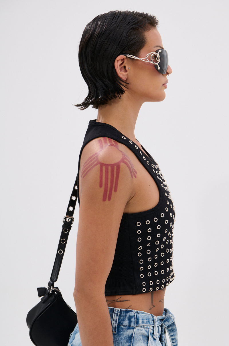 A person with short dark hair wears large decorative sunglasses, blue jeans, and the black JANE GROMMET TANK TOP with metallic studs. They carry a black shoulder bag and have a red abstract tattoo on their upper arm against a plain white background. 3