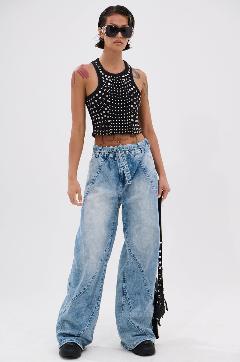 A person with short hair, sunglasses, and visible tattoos wears the JANE GROMMET TANK TOP and loose, light-wash wide-leg jeans, holding a black strap accessory against a plain white background. 6