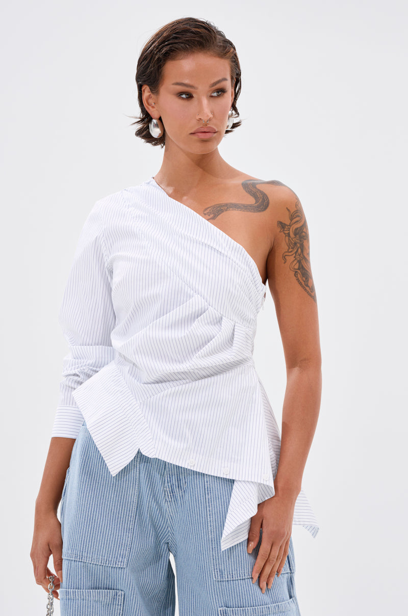 A person with short dark hair and earrings wears the ASSIA ONE SHOULDER BLOUSE and blue pinstripe cargo pants, revealing a snake tattoo on their shoulder as they stand against a plain, light background.