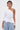 A woman with short, dark hair and a shoulder tattoo wears the ASSIA ONE SHOULDER BLOUSE—white, pinstriped, with an asymmetrical neckline—paired with light blue pinstriped cargo pants, standing against a plain light background.