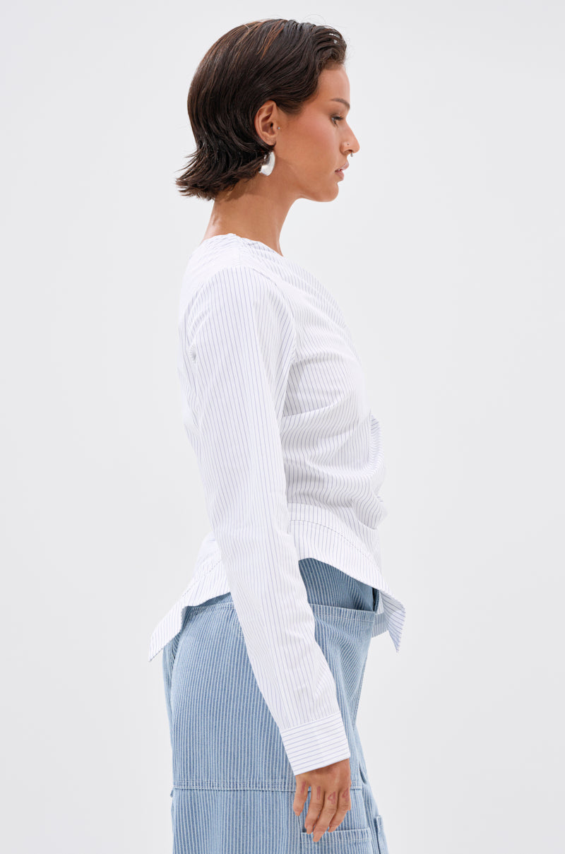 A woman with short dark hair models the ASSIA ONE SHOULDER BLOUSE, standing in profile against a plain light background.