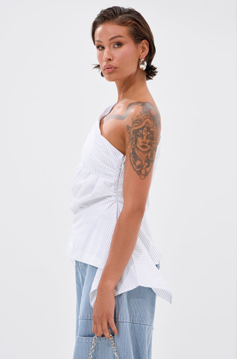 A person with short dark hair, silver earrings, and a large upper arm tattoo models the white ASSIA ONE SHOULDER BLOUSE with blue pinstriped pants against a plain white background.