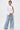 A person stands confidently in the ASSIA ONE SHOULDER BLOUSE with an asymmetrical neckline, paired with light blue wide-leg jeans, white sneakers, and a metallic handbag against a plain white background.