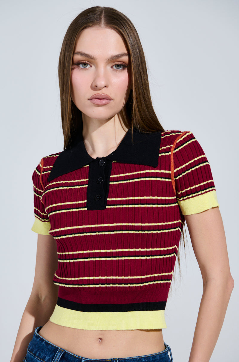 A person with long brown hair is wearing the SCHOOL GIRL VIBES POLO, featuring a cropped silhouette in red, yellow, and black stripes. This ribbed knit top has short sleeves, a large black collar, and front buttons. They're against a plain background, exuding school girl vibes. 0