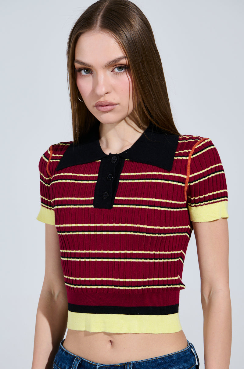 A person dons the SCHOOL GIRL VIBES POLO, a short-sleeved, ribbed knit top with red and yellow stripes and a black collar. The buttoned front adds charm, perfectly matched with blue jeans against a plain, light-colored backdrop. 1