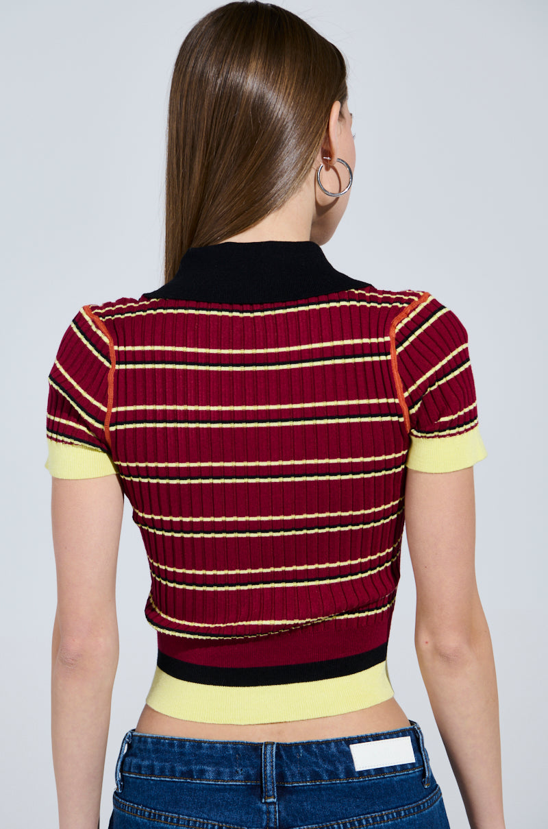 A person with long brown hair and hoop earrings is seen from behind, wearing a SCHOOL GIRL VIBES POLO—a short-sleeve ribbed knit top with red, yellow, and black stripes—and blue jeans. 3