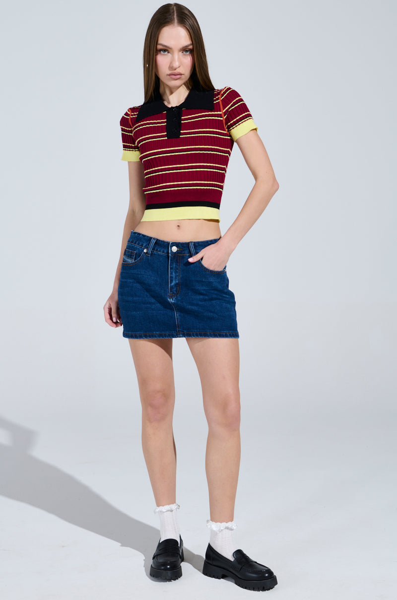With long hair against a plain backdrop, they embody school girl vibes in the SCHOOL GIRL VIBES POLO—a striped red and yellow top, blue denim skirt, white socks, and black slip-on shoes. The cropped silhouette adds playfulness as one hand sits in their pocket and the other by their side. 4
