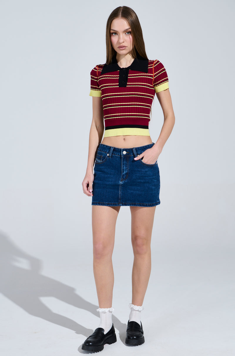A person with long hair stands against a plain background, embodying school girl vibes in the SCHOOL GIRL VIBES POLO—a red and yellow striped shirt with a black ribbed knit collar. A denim skirt, white socks, and black shoes complete the look as one hand rests in the skirt pocket. 5