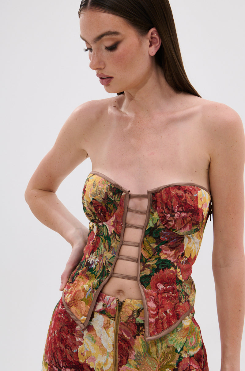 A woman with long brown hair wears the LILY BROCADE CORSET, a strapless corset with a floral tapestry design, paired with matching high-waisted bottoms. She poses against a plain white background, gazing downward.