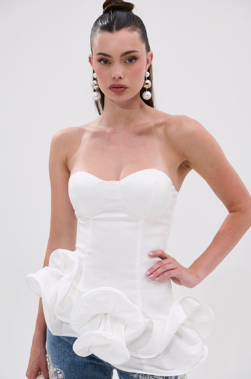 A woman with long brown hair in a high ponytail wears large pearl earrings and THE MORE YOU KNOW RUFFLE DETAIL CORSET TOP with a strapless sweetheart neckline and peplum hem, paired with blue jeans against a plain light background.