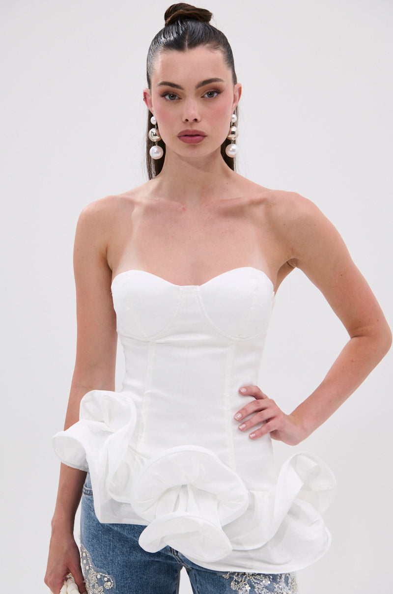 A woman with long dark hair in a ponytail wears statement pearl earrings, blue jeans with silver embellishments, and THE MORE YOU KNOW RUFFLE DETAIL CORSET TOP, standing against a plain white background.
