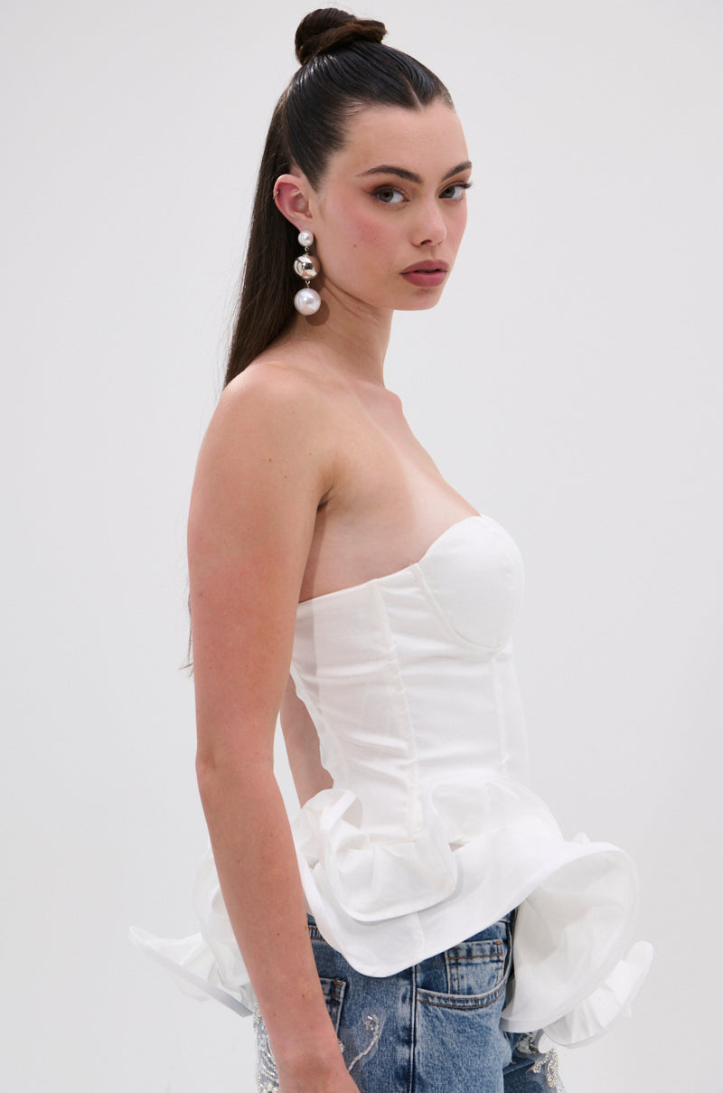 A woman with long dark hair in a high bun wears large pearl earrings and the THE MORE YOU KNOW RUFFLE DETAIL CORSET TOP, paired with distressed blue jeans, posing against a plain light background.